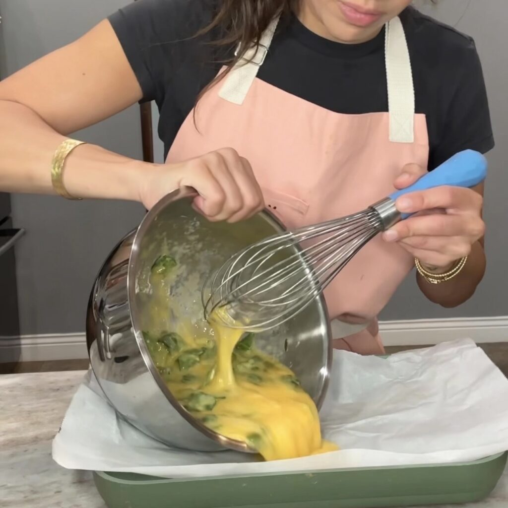 Pouring spinach egg mixture onto a parchment-lined sheet pan for high protein breakfast burritos meal prep